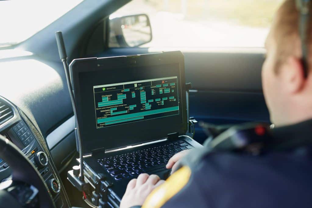 Database, laptop and a police officer in a car for security, urban law and safety data while working. Screen, programming and male protection professional with a computer to hack system in transport. Alexandria Police Department benefits