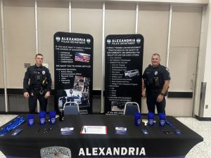 Alexandria Police Department recruitment booth featuring two uniformed officers standing behind a display table with brochures, branded cups, and banners promoting career opportunities and benefits — focus key phrase: Alexandria Police Department.